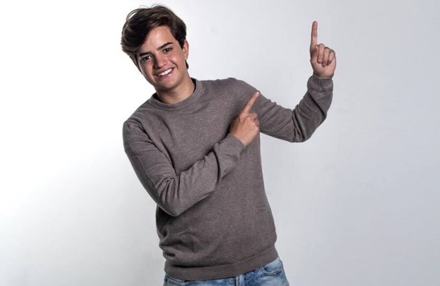 Jovem sorridente, com moletom e jeans, aponta com dois dedos para o lado e para cima.