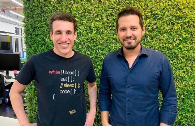 Dois homens sorrindo, um com camiseta de programação, à frente de parede de folhagem verde.