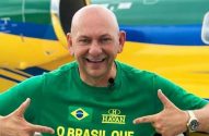 Homem careca sorrindo, apontando para camisa verde com a bandeira do Brasil. Fundo azul e amarelo.