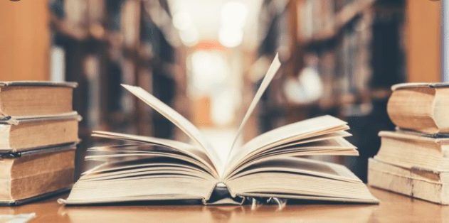 Um livro aberto sobre mesa com pilhas de livros ao lado. Fundo de biblioteca desfocado.