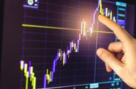 Mão apontando para gráfico financeiro em tela, mostrando tendência de alta.