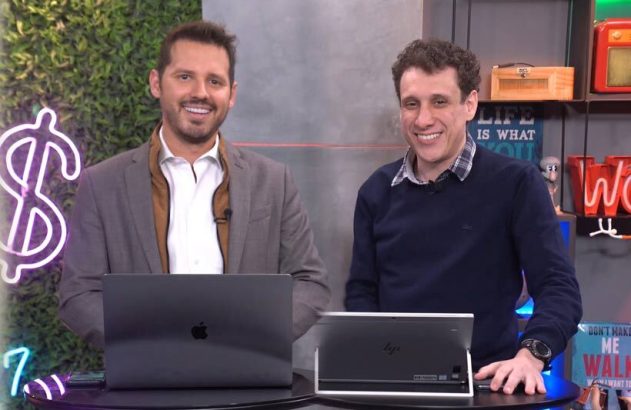 Dois homens sorrindo sentados em mesa com laptops em um estúdio.