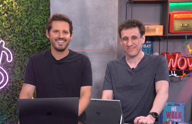 Dois homens sorrindo olham para a câmera em um estúdio com laptops à frente.