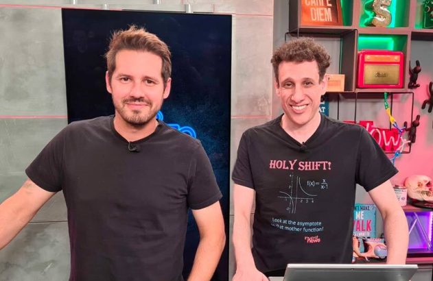 Dois homens em camisetas pretas posam lado a lado em um estúdio. Um deles tem barba, o outro cabelo cacheado.