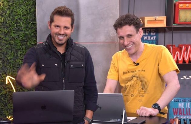 Dois homens sorrindo, um acenando, atrás de laptops em um estúdio.
