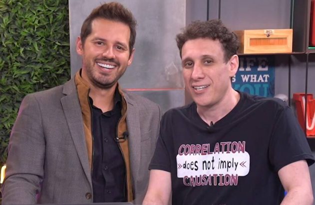 Dois homens sorrindo. Um de blazer, outro com camiseta 'Correlation does not imply causation'. Fundo de estúdio.