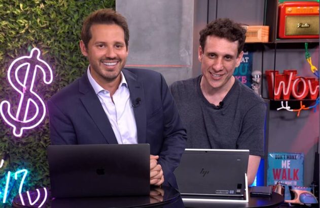 Dois homens sorrindo, sentados com laptops, apresentando em um estúdio com letreiros neon ao fundo.