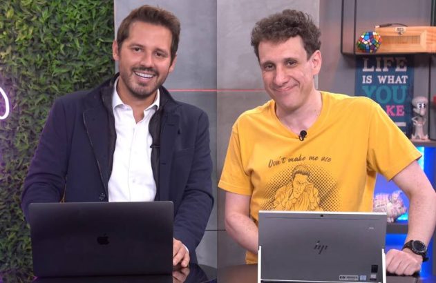Dois apresentadores sorriem enquanto trabalham em laptops em estúdio.