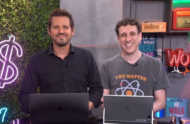 Dois homens sorrindo, em pé, com laptops. Fundo de estúdio com luzes neon (cifrão, WOW) e prateleiras.