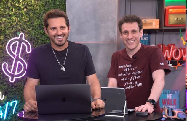 Dois homens sorrindo em estúdio, sentados a uma mesa com laptop e tablet. Um neon de cifrão no fundo.