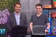 Dois homens sorridentes, com laptops em mesas pretas, olhando para a câmera. Estão em um estúdio com fundo de plantas e prateleiras.