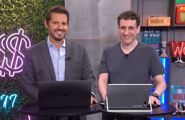 Dois homens sorridentes, com laptops em mesas pretas, olhando para a câmera. Estão em um estúdio com fundo de plantas e prateleiras.