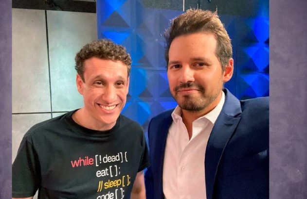 Dois homens sorridentes, um de camisa preta com código e outro de terno azul, posam para foto.