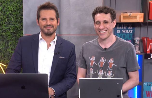 Dois homens sorrindo: um de terno, outro de camiseta, posam com laptops em um estúdio.