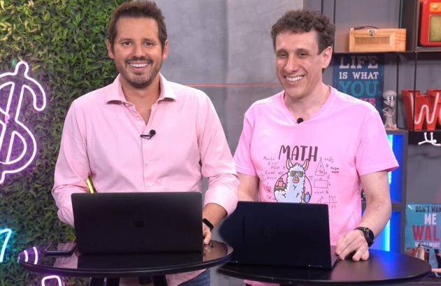Dois homens em camisas rosa sorriem para a câmera, atrás de laptops com fundo de cifrão neon.