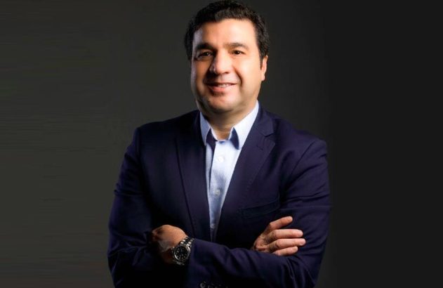 Retrato profissional: homem sorridente de terno azul e camisa clara, com braços cruzados.