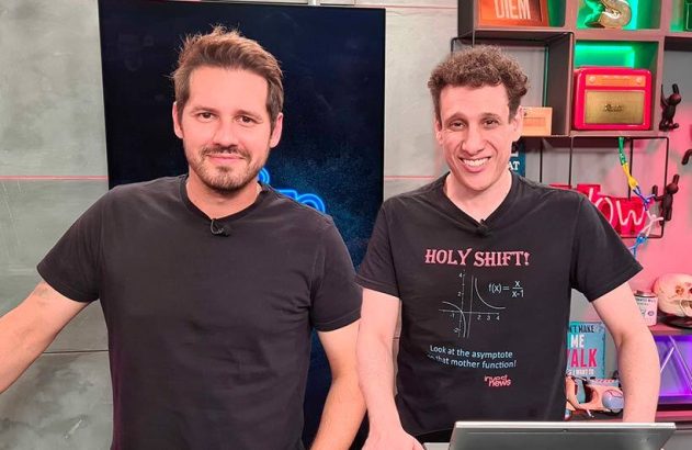 Dois homens sorrindo em estúdio de TV, um com camiseta de piada de matemática.