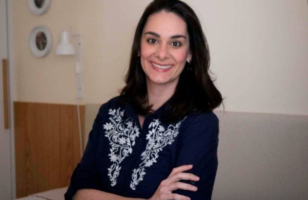 Mulher sorridente de cabelo escuro, camisa azul bordada com braços cruzados, olhando para a câmera.