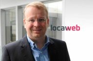 Homem de óculos, sorrindo, em traje profissional, com logo Locaweb ao fundo.