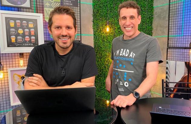 Dois homens sorrindo para a câmera em estúdio, um sentado com notebook e outro em pé.