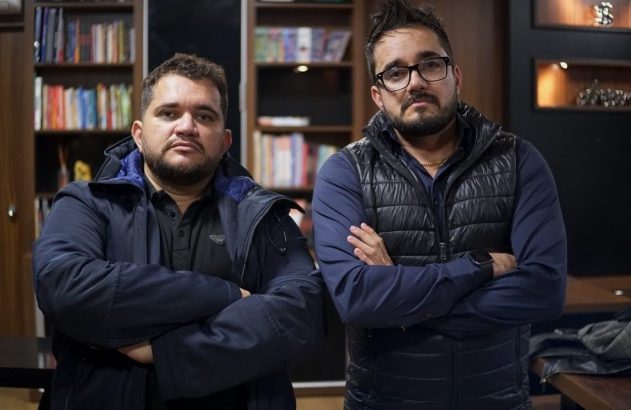 Dois homens de braços cruzados posam em um escritório com estantes de livros.