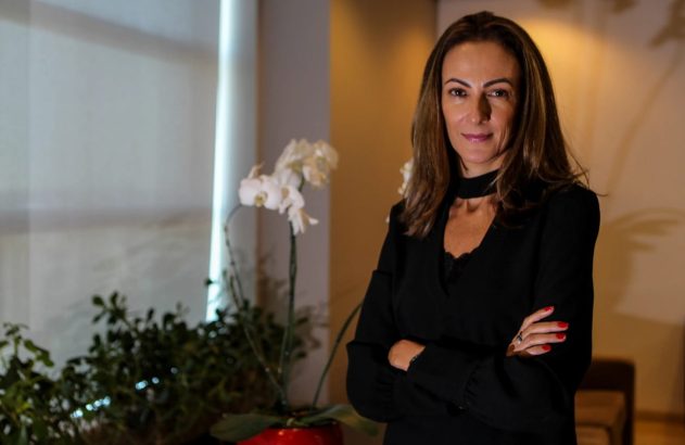 Retrato de mulher de cabelos castanhos, blusa preta, braços cruzados, sorrindo em ambiente interno.