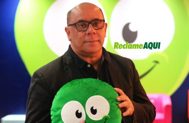 Homem de óculos segura mascote verde redondo. Logo 'Reclame AQUI' visível ao fundo.