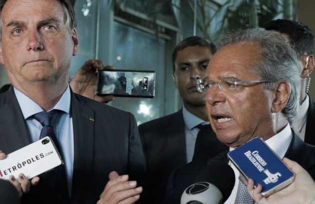 Jair Bolsonaro e Paulo Guedes em coletiva de imprensa. Guedes fala com microfones à frente; Bolsonaro atento.