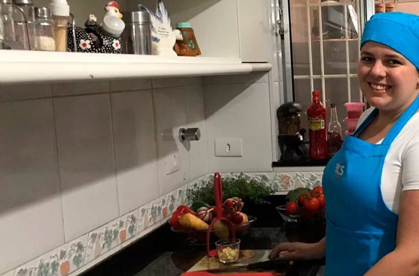 Cozinheira sorridente de avental e touca azuis em uma cozinha, com alimentos na bancada.