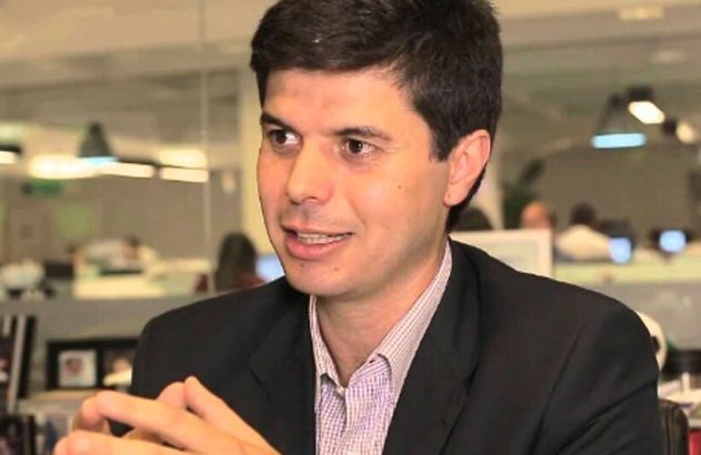 Homem de terno e camisa listrada fala atentamente em escritório, mãos juntas.