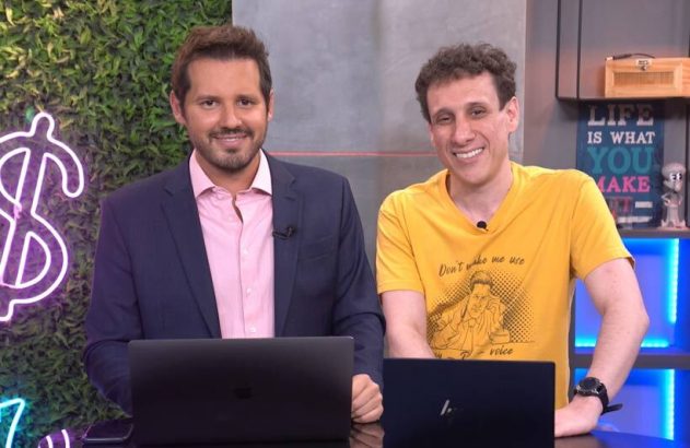 Dois homens sorrindo, um de terno e outro de camiseta amarela, atrás de laptops, em um estúdio.