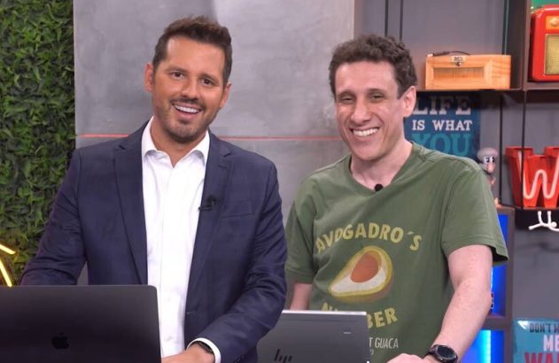 Dois apresentadores sorridentes, um de terno e outro de camiseta, atrás de bancadas com laptops.