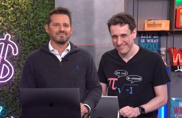 Dois homens sorrindo em um cenário de estúdio. O da direita veste camiseta com tema de matemática. Há laptops e decoração.