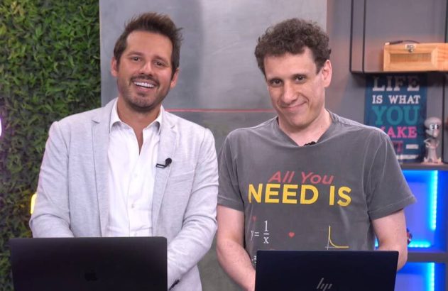 Dois homens sorrindo em estúdio com laptops. Um usa jaqueta clara, outro camiseta escura.