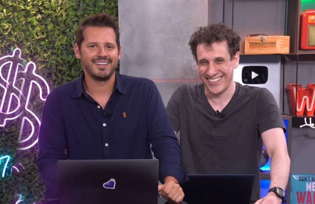 Dois homens sorrindo em um estúdio, à frente de laptops. Fundo com neon verde e prêmios.