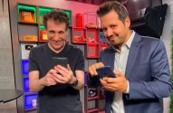 Dois homens sorridentes seguram smartphones e olham para a câmera em um ambiente interno colorido.