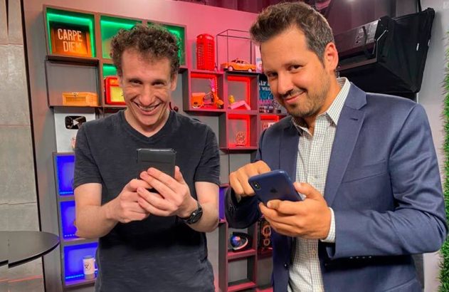 Dois homens sorridentes seguram smartphones e olham para a câmera em um ambiente interno colorido.