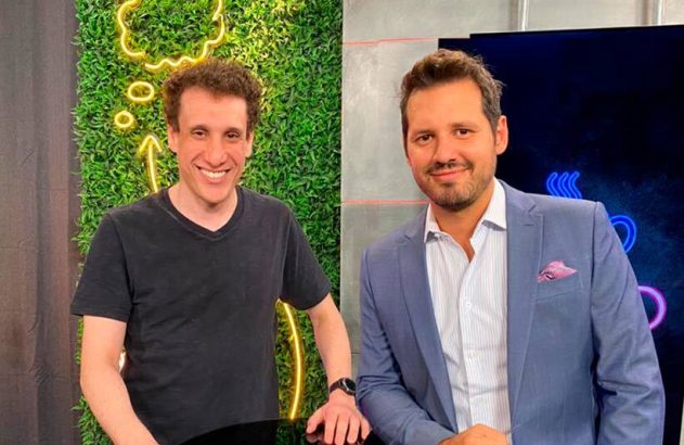 Dois homens sorridentes posam em estúdio. Um usa camiseta preta, o outro um paletó azul. Fundo com neon.