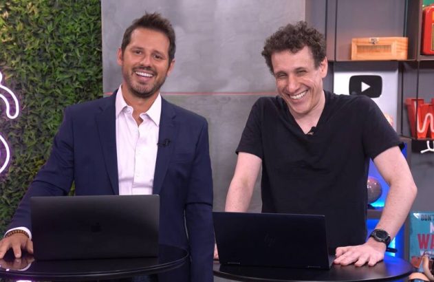 Dois homens sorridentes com laptops em estúdio. Um de terno azul, outro de camiseta preta.