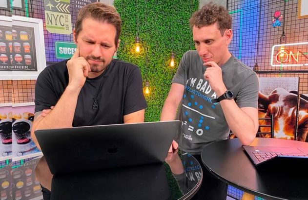 Dois homens, um sentado e outro em pé, olham atentamente para um notebook.