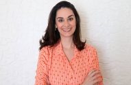 Mulher sorrindo, cabelos escuros, camisa pêssego de flechas, braços cruzados, fundo branco texturizado.