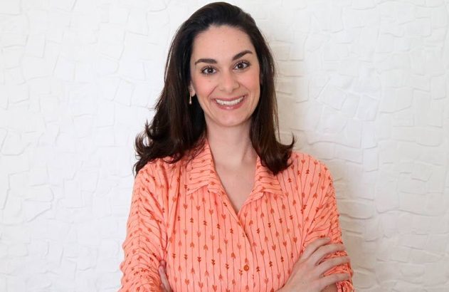 Mulher sorrindo, cabelos escuros, camisa pêssego de flechas, braços cruzados, fundo branco texturizado.