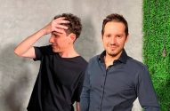 Dois homens. Um à esquerda com a mão na cabeça e olhando para o lado; outro à direita sorrindo para a câmera.