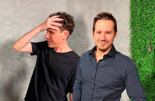 Dois homens. Um à esquerda com a mão na cabeça e olhando para o lado; outro à direita sorrindo para a câmera.