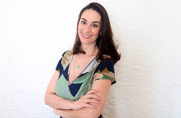 Mulher sorridente de pé, braços cruzados, cabelo escuro e vestido estampado, fundo branco.