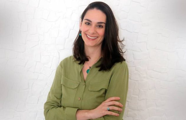 Mulher sorrindo, de camisa verde e braços cruzados, em frente a uma parede branca texturizada.