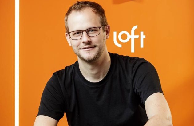 Homem sorridente de óculos e camiseta preta, em fundo laranja com o logo da Loft.