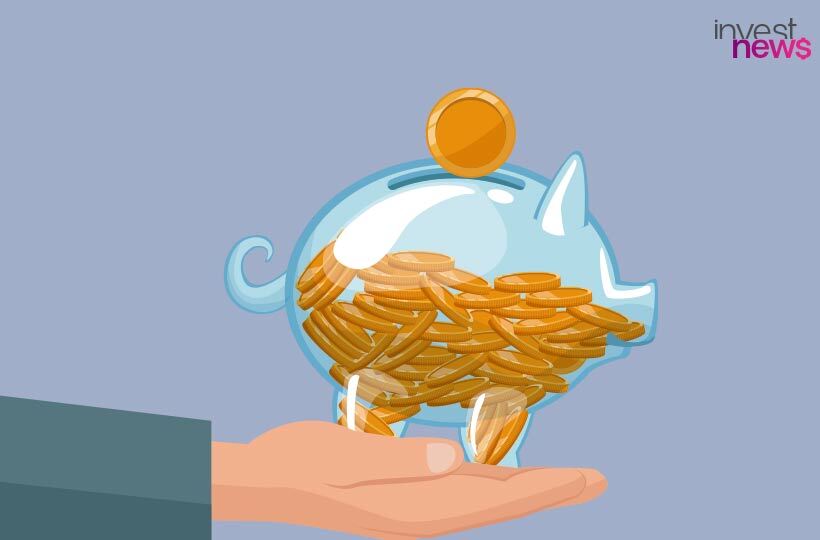 Mão segura cofrinho azul transparente cheio de moedas de ouro, com uma moeda sendo depositada.