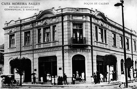 Foto antiga P&B: Edifício da Casa Moreira Salles em Poços de Caldas, estabelecimento comercial e bancário, com pessoas e carro.