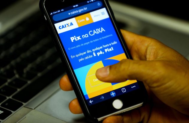 Mão segurando smartphone com tela mostrando "Pix na CAIXA" e caixa.gov.br. Dedo toca na tela.